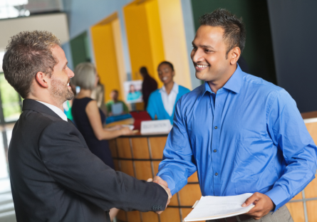 Two men in professional attire shake hands at a job fair. One of the men holds a resume to give to the potential employer.