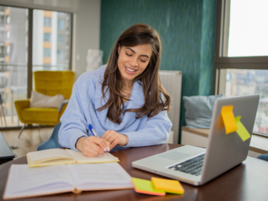 Woman writing in notebook while working on laptop