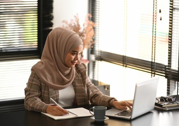 Woman reading laptop screen and writing in notebook